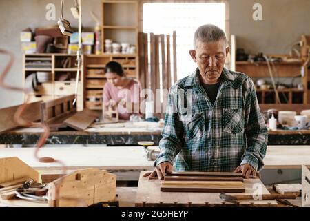 Serious pensive senior carpenter looking at pieces of walnut and maple wood that he wants to fuse together for making edge grain cutting board Stock Photo