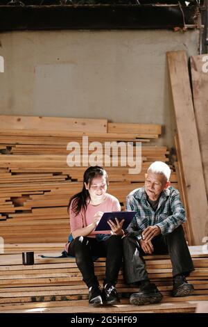 Proud smiling senior carpenter looking at his granddaughter sketching furniture item for customer Stock Photo