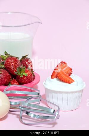 Cupcake with cream and strawberry slices, beaters from the mixer, egg, milk on a pink background. Selective focus. Stock Photo
