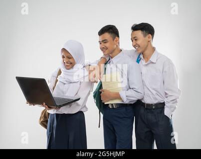 Three teenagers in junior high school uniforms smiling using a laptop computer together while carrying a backpack and a book Stock Photo