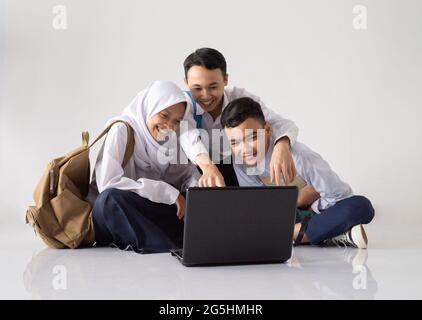 smiling three teenagers in junior high school uniforms sitting on the floor while using a laptop together Stock Photo