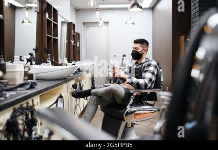 Young man haidresser using smartphone in barber shop, coronavirus and new normal concept. Stock Photo