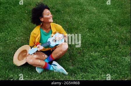 Rastafarian fun woman is passionate about playing the ukulele sitting on the green lawn, she enjoys summer and music Stock Photo