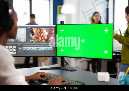 African video editor with headset editing footage using pc with greenscreen, chroma key isolated display sitting in creative agency. Videographer in digital multimedia company processing movie Stock Photo