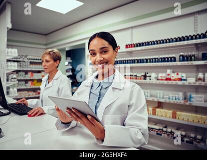 Portrait of smiling young female pharmacist wearing labcoat holding digital tablet with senior colleague working in background on computer Stock Photo