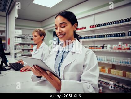 Happy woman working in medical store wearing labcoat using digital tablet with colleague working in background Stock Photo