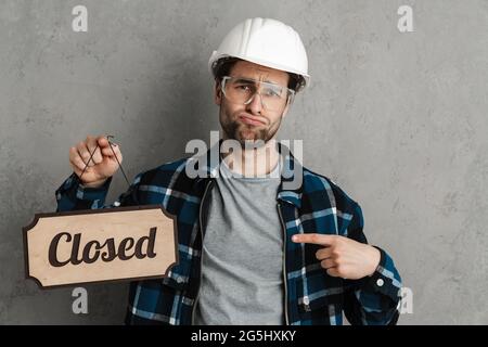 Unhappy builder man holding and pointing finger at closed sign board isolated over grey wall Stock Photo