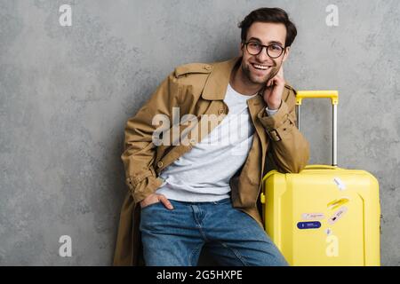 Attractive happy young man wearing coat standing with suitcase ready for travel isolated over gray Stock Photo