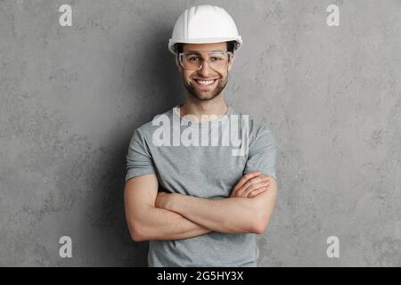 Joyful builder man in helmet and goggles smiling and looking at camera isolated over grey wall Stock Photo