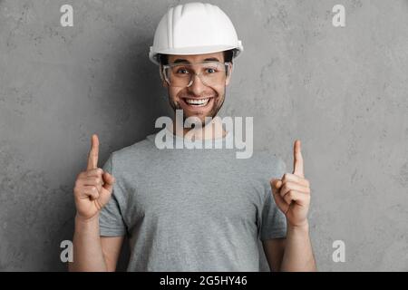 Joyful builder man in helmet and goggles smiling and pointing finger upward isolated over grey wall Stock Photo