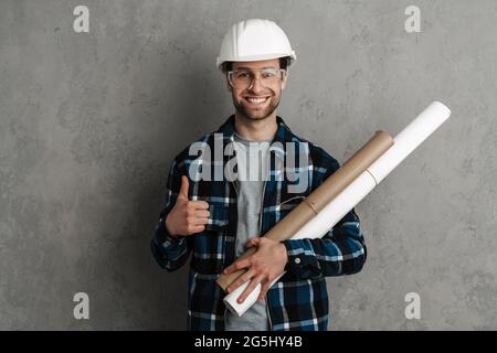 Joyful builder man in helmet and goggles showing thumb up isolated over grey wall Stock Photo