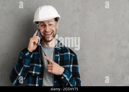 Joyful builder man talking on cellphone and pointing finger aside isolated over grey wall Stock Photo