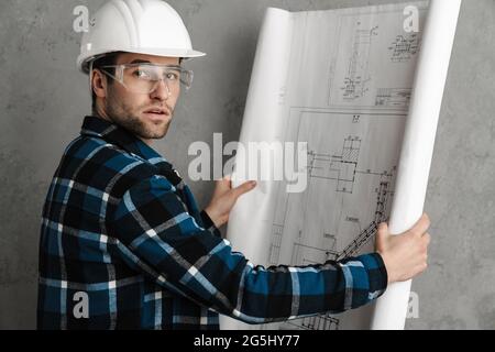 Young handsome civil engineer in hardhat holding blueprints standing isolated over gray background Stock Photo