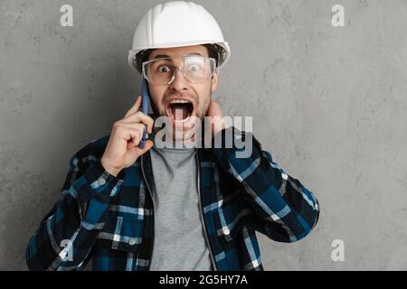 Shocked builder man in helmet and goggles talking on mobile phone isolated over grey wall Stock Photo