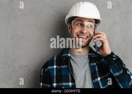 Joyful builder man in helmet and goggles talking on mobile phone isolated over grey wall Stock Photo