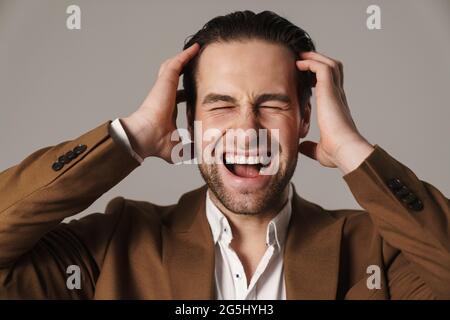 Unshaven irritated man in jacket screaming while holding his head isolated over grey background Stock Photo