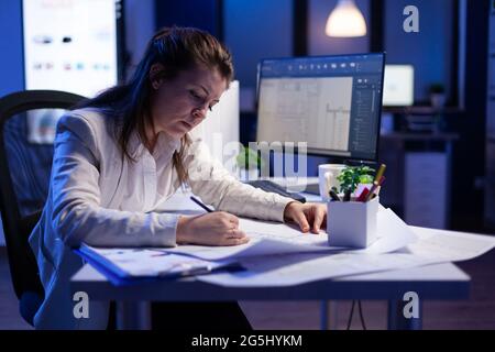 Overworked woman architect checking and matching blueprints sitting at office desk in front of computer. Perfectionist designer using arhitecture blueprints of buildings creating industrial prototype Stock Photo