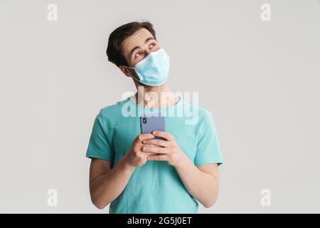 Joyful handsome guy in protective mask using mobile phone isolated over white wall Stock Photo