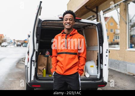 Portrait of smiling essential service man standing with hands in pockets against delivery van Stock Photo