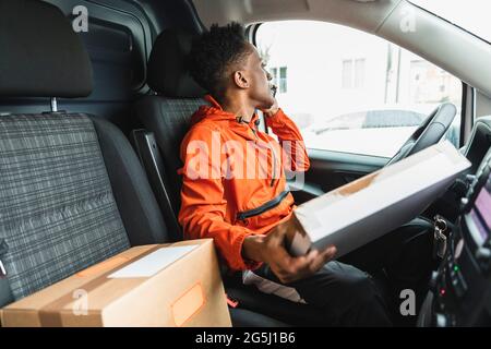 Delivery man holding package talking on mobile phone while looking away through van window Stock Photo