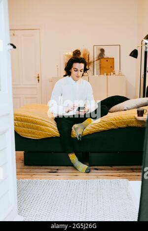 Full length of man using smart phone while sitting on bed at home Stock Photo