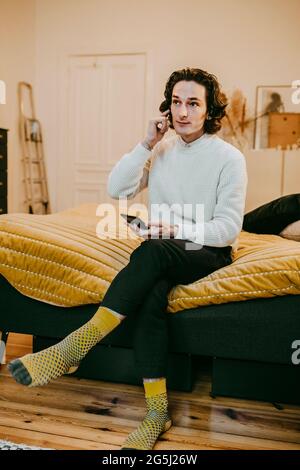 Man wearing in ear-headphones sitting with mobile phone on bed at home Stock Photo