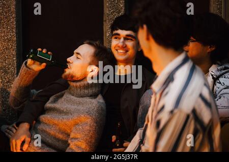 Young man drinking beer while chilling with male friends in balcony Stock Photo