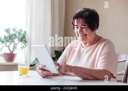 An elderly woman uses a tablet at home and watches content on social networks - An elderly person knows how to use modern technologies Stock Photo