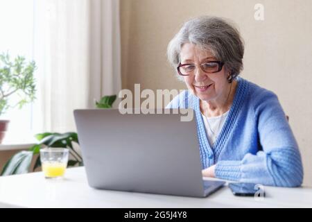 Elderly business woman with glasses working in her home office looking for information or chatting on a social network - Pensioner uses a laptop to vi Stock Photo