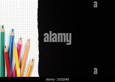 Colored pencils on white squared torn sheet of paper over black school chalkboard blackboard. Back to school, learning concept. Top view, copy space Stock Photo