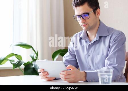 A young man uses a tablet at home while sitting at a table - A Millennial with glasses is working on a new project - A man uses a modern gadget and lo Stock Photo