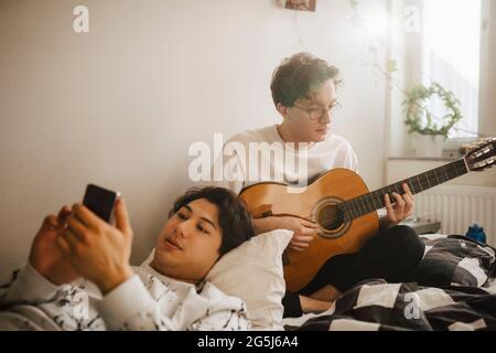 Young man using smart phone while male friend playing guitar at home Stock Photo