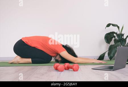 Girl at home on a sports mat doing a workout - Young woman doing sports exercises at home using a video call on a laptop - Girl makes relaxing body mo Stock Photo
