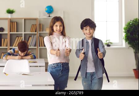Boy and girl elementary school students show thumbs up standing in school classroom. Stock Photo