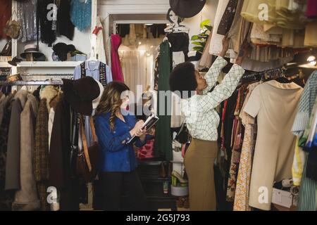 Female employee arranging clothes in rack while employee making notes at store Stock Photo