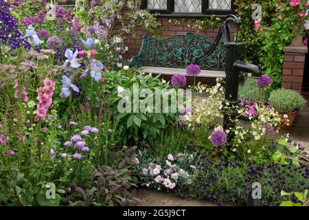 The Cottage Garden' featuring a water pump and wrought iron and wooden seat surrounded by Allium 'Purple Sensation',  and Foxgloves. Stock Photo