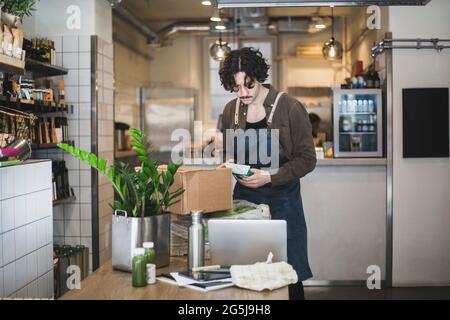 Young male employee working on laptop while holding food package in store Stock Photo