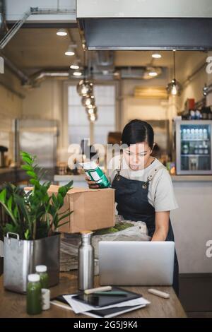 Female employee working laptop while holding food package at store Stock Photo