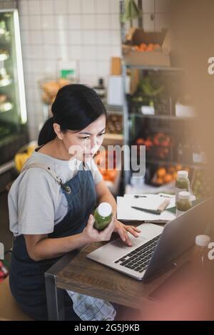 Asian female owner using laptop while juice bottle in store Stock Photo