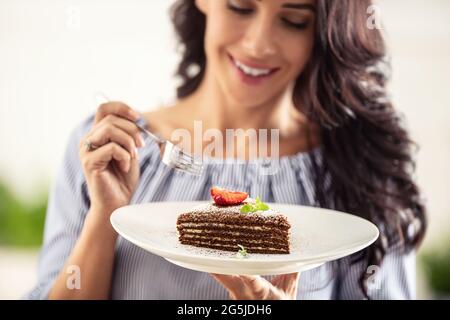 Layered brown adn white cake with strawberry and mint leaf on top held by a woman with a fork. Stock Photo