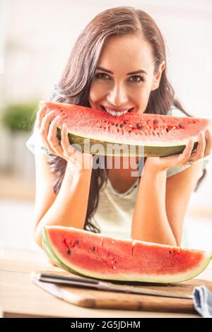 Good-looking girl bites into the the quarter of a watermelon in the kitchen, smiling. Stock Photo