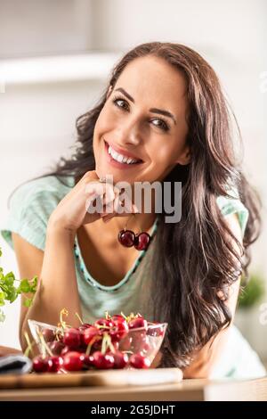 Smiling woman picks two cherries from a seethrough bowl indoors. Stock Photo