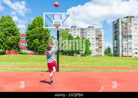 Attractive young boy shooting ball to the hoop at playground.cute young boy plays basketball on the playground in the summer warm day. Stock Photo