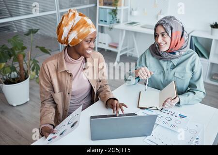 High angle portrait of two ethnic young women wearing headscarf while enjoying work together in office, copy space Stock Photo