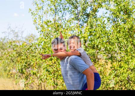 Defocused brother giving sister ride on back. Portrait of happy girl with funny horns on man shoulders, piggyback. Family playing outside. Summer Stock Photo