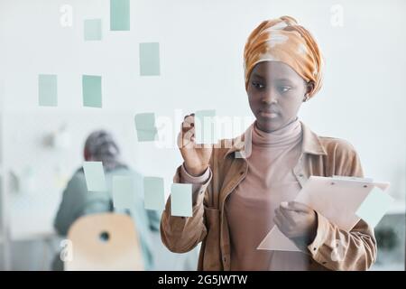 Waist up portrait of modern African-American woman working in office and placing sticker notes on glass wall, copy space Stock Photo