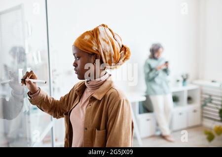 Side view portrait of modern African-American woman writing notes on glass wall while working in office, copy space Stock Photo