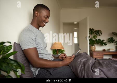 Smiling African man using a tablet and drinking coffee in bed Stock Photo