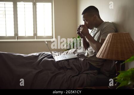 Young African man using a laptop and drinking coffee in bed Stock Photo