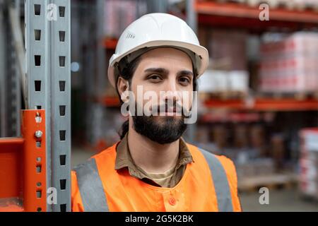 Confident worker of warehouse looking at you while standing by rack Stock Photo